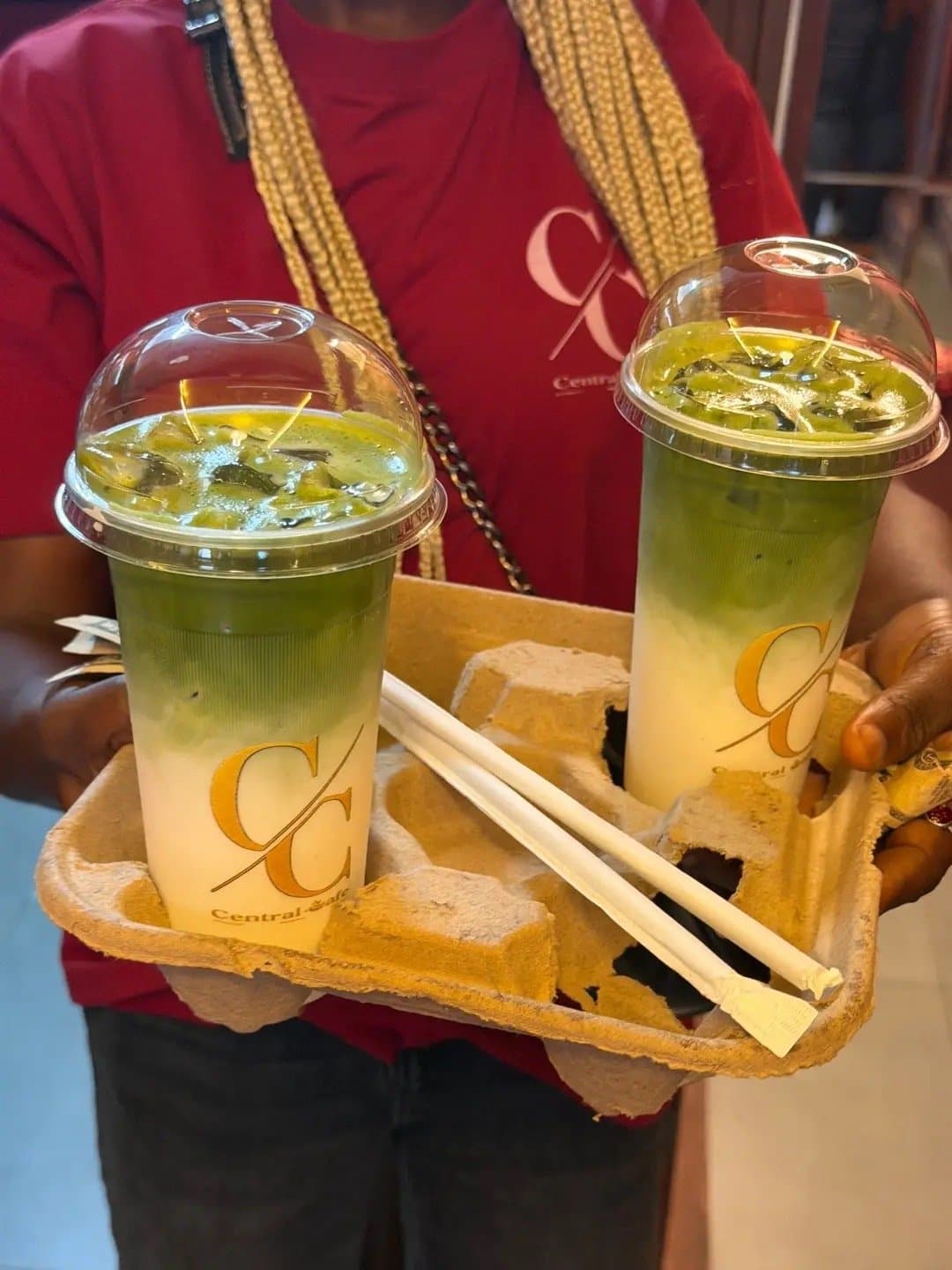 Two layered matcha drinks in a carry tray, held out by a barista in a Central Cafè shirt